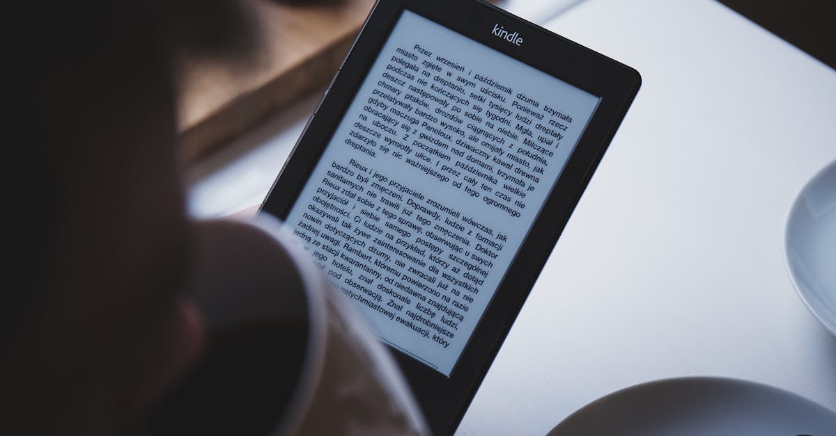 A person reads on a Kindle e-reader while holding a cup of coffee, indoors.